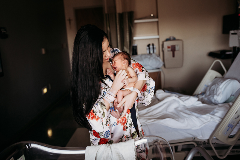 Fresh 48 Photography, A new mother holds her crying baby in the hospital room as she tends to it