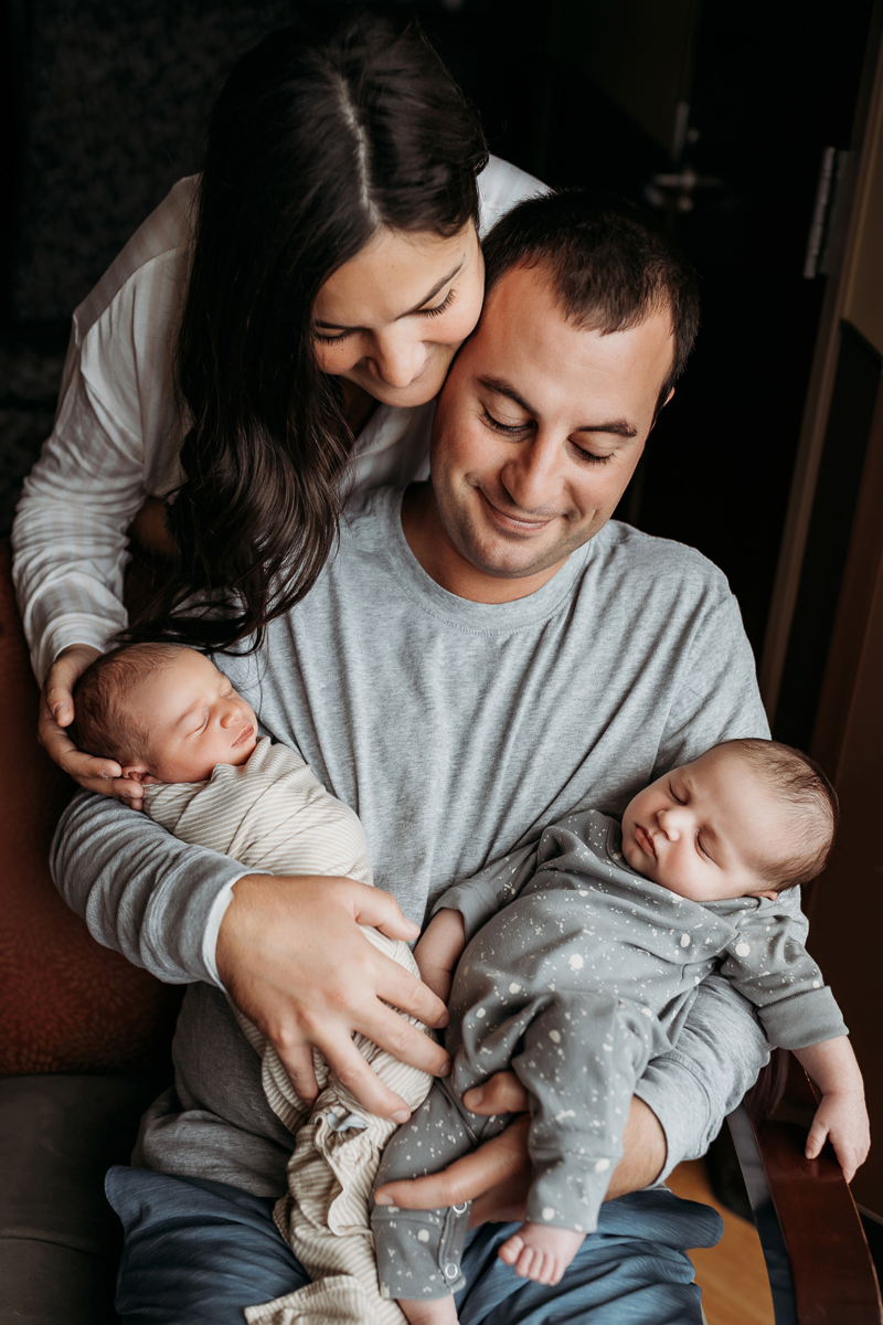 Fresh 48 Photography, mom leans over dads shoulder as he holds two newborn babies