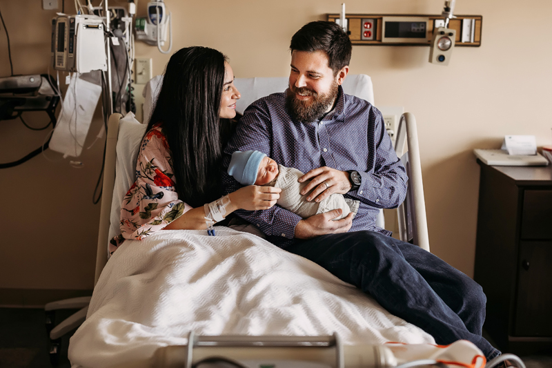 Fresh 48 Photography, dad sits with mom in her hospital bed as they hold their newly delivered baby