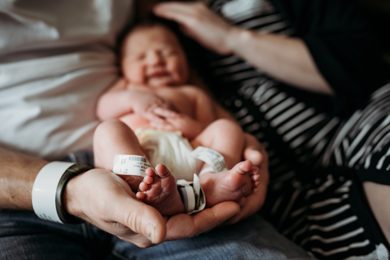 Fresh 48 Photography, a father holds his newborn baby, the baby's feet sit dwarfed in dad's large hands, mom is nearby too