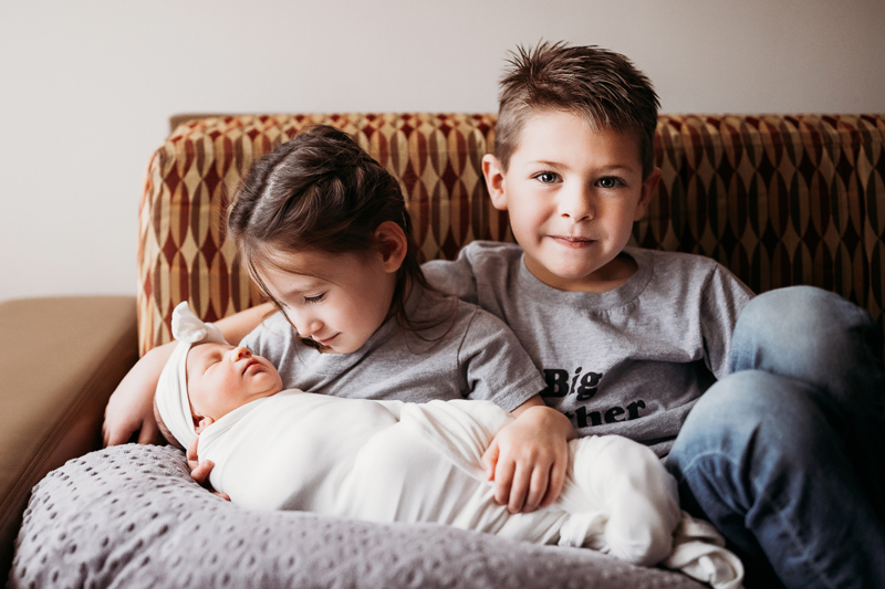 Fresh 48 Photography, a little boy sits on the couch with his arm wrapped around sister, the sister looks down at her newborn sibling in her arms