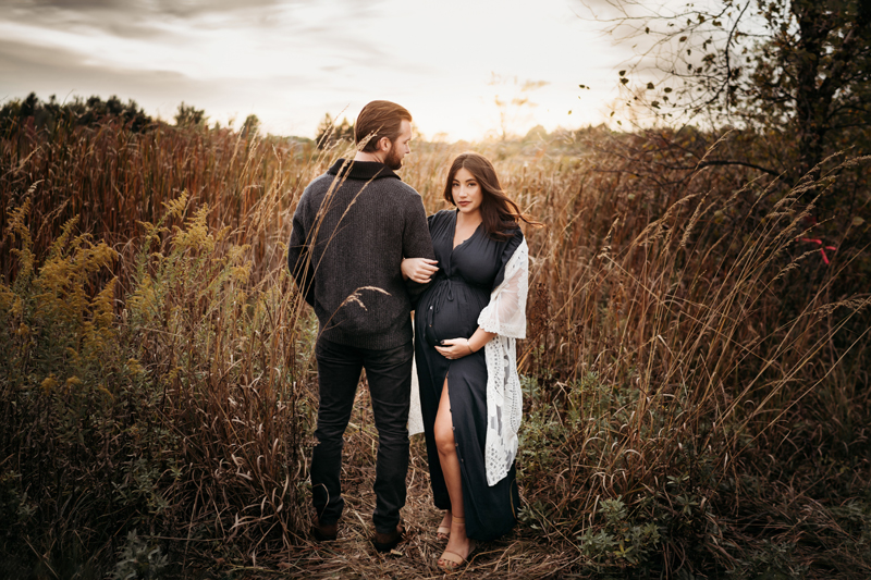 Maternity Photography, an expecting woman holds her belly and onto her man outdoors, he is faced the other direction looking back at his wife