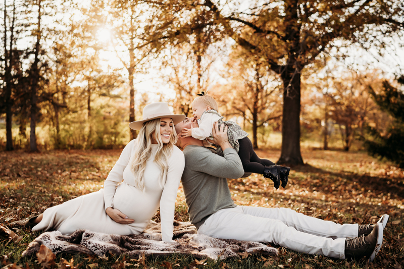 Maternity Photography, a man and his expecting partner sit on a blanket outdoors in the park. he holds their young daughter