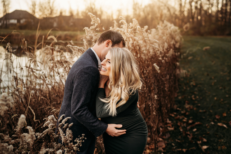 Maternity Photography, a man and woman embrace near a lake, she is expecting, they both smile.