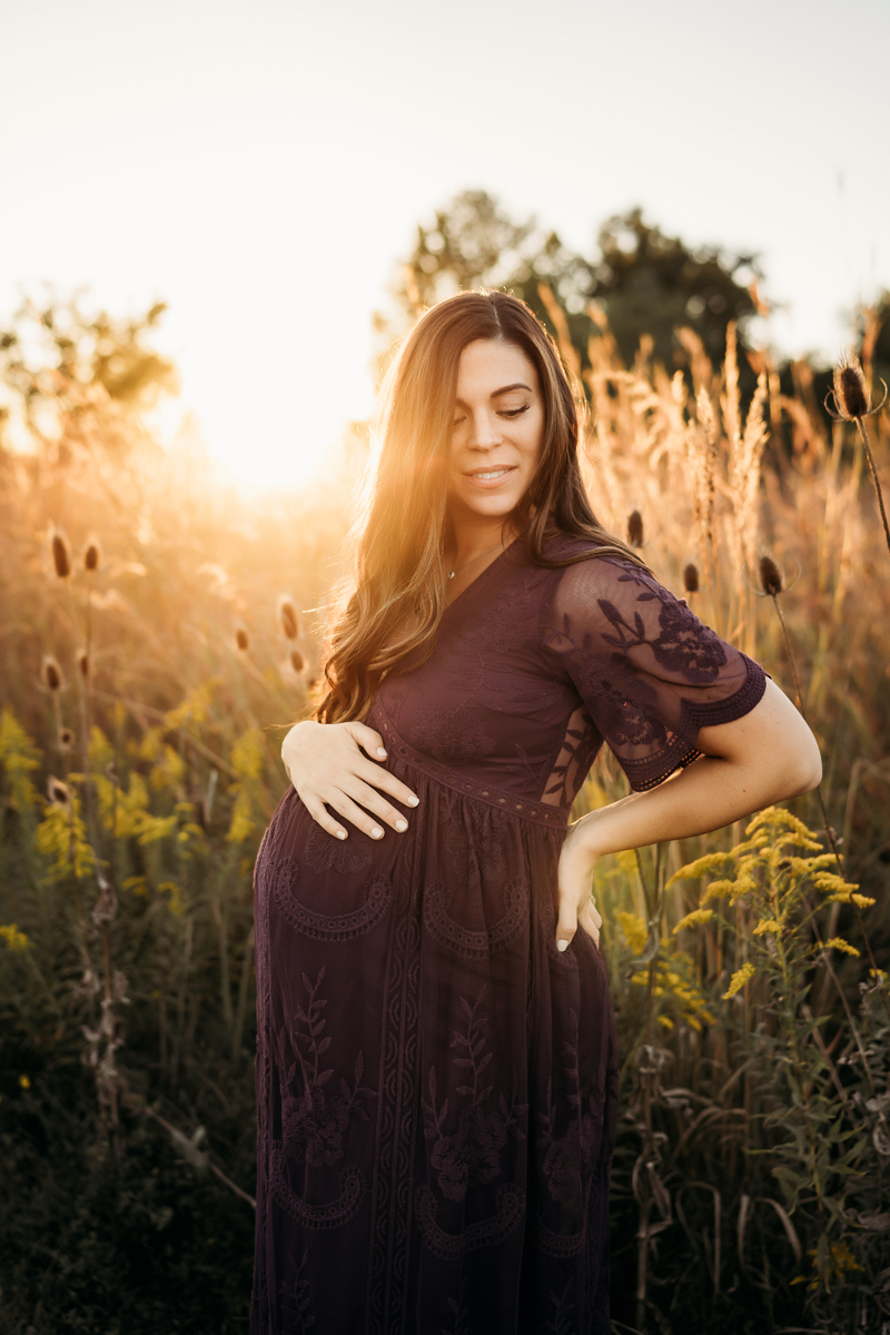 Maternity Photography, a woman in a maroon dress lays her hand on her pregnant belly in a field