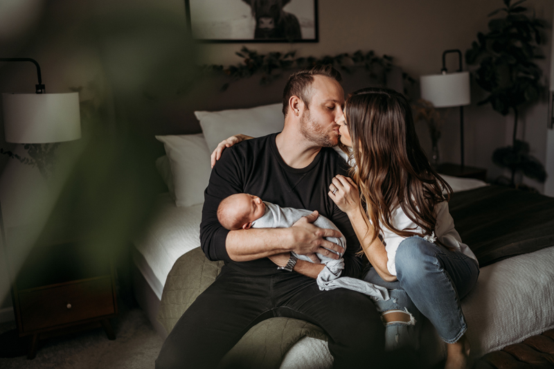 Newborn Photography, woman kisses man as he holds their newborn baby daughter within their bedroom